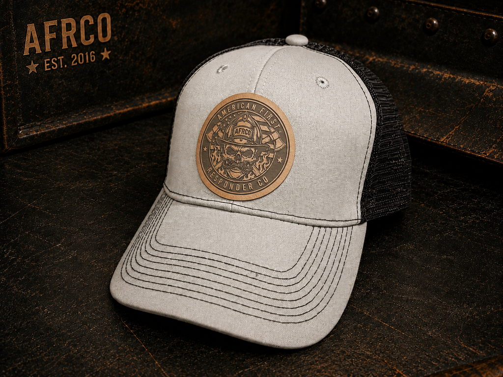 A light gray heather trucker cap with a black mesh back, featuring a circular bronze-toned metal patch on the front with a firefighter skull design and the words 'American Fire Service' and 'LIFELONG FIRE DEPT.'.