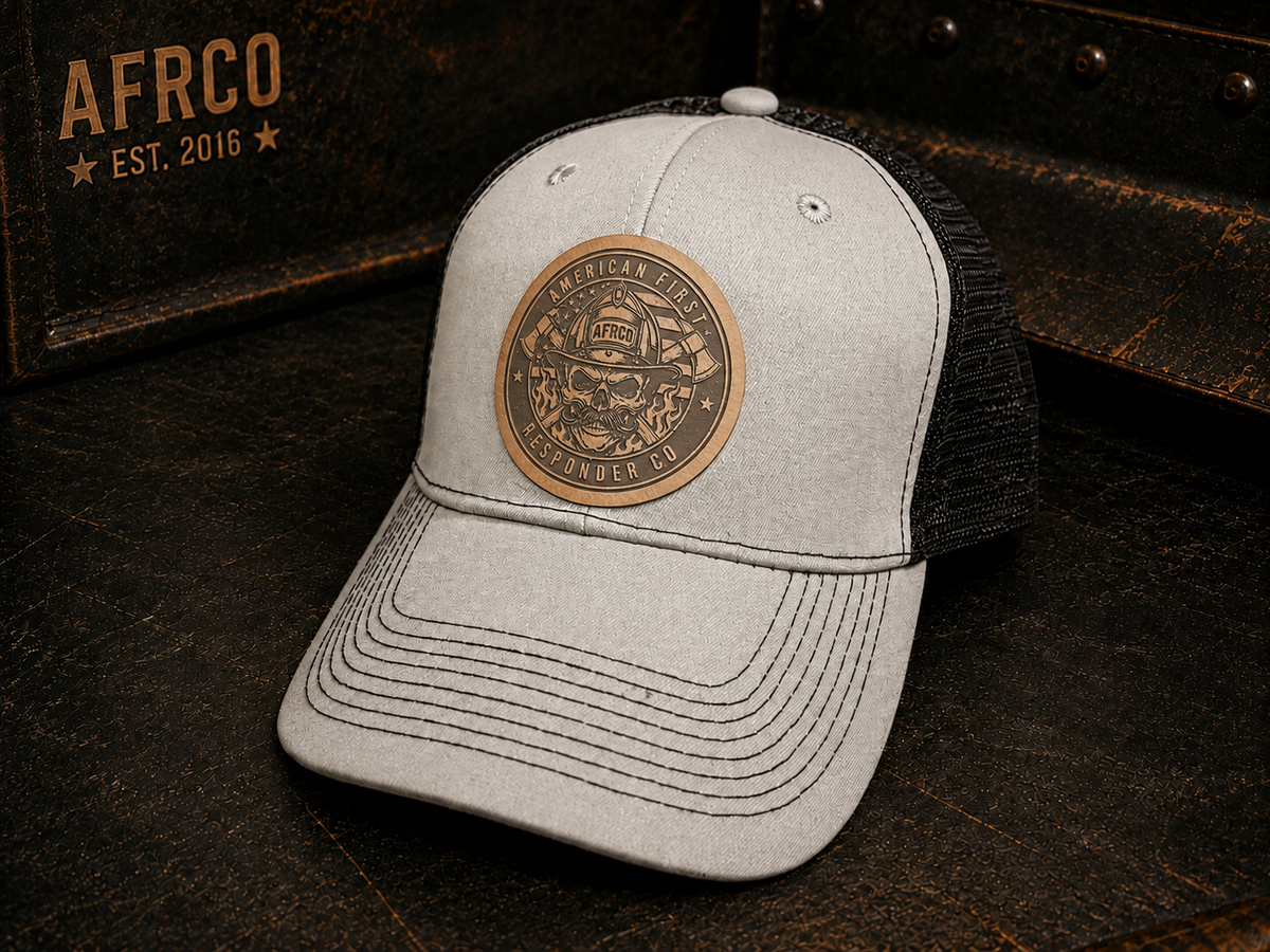 A light gray heather trucker cap with a black mesh back, featuring a circular bronze-toned metal patch on the front with a firefighter skull design and the words 'American Fire Service' and 'LIFELONG FIRE DEPT.'.