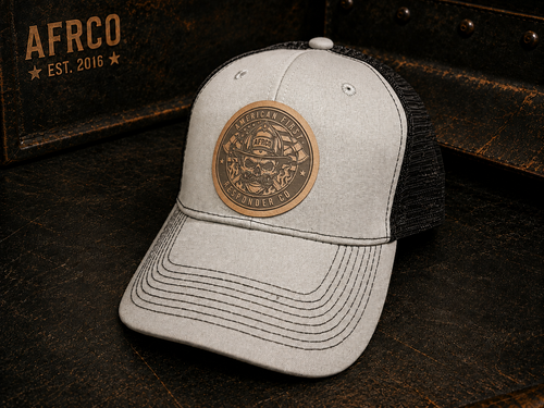A light gray heather trucker cap with a black mesh back, featuring a circular bronze-toned metal patch on the front with a firefighter skull design and the words 'American Fire Service' and 'LIFELONG FIRE DEPT.'.